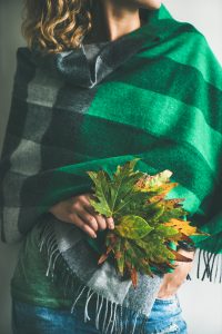 Woman in warm woolen green check scarf or blanket and blue jeans with Autumn fallen leaves in her hands. Fall cosy mood lifestyle concept